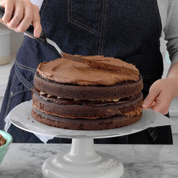 Person frosting a three-layer chocolate cake on a white pedestal stand. The cake has chocolate filling between the layers. They are spreading icing on the top with a spatula. The background includes a countertop and kitchen items.