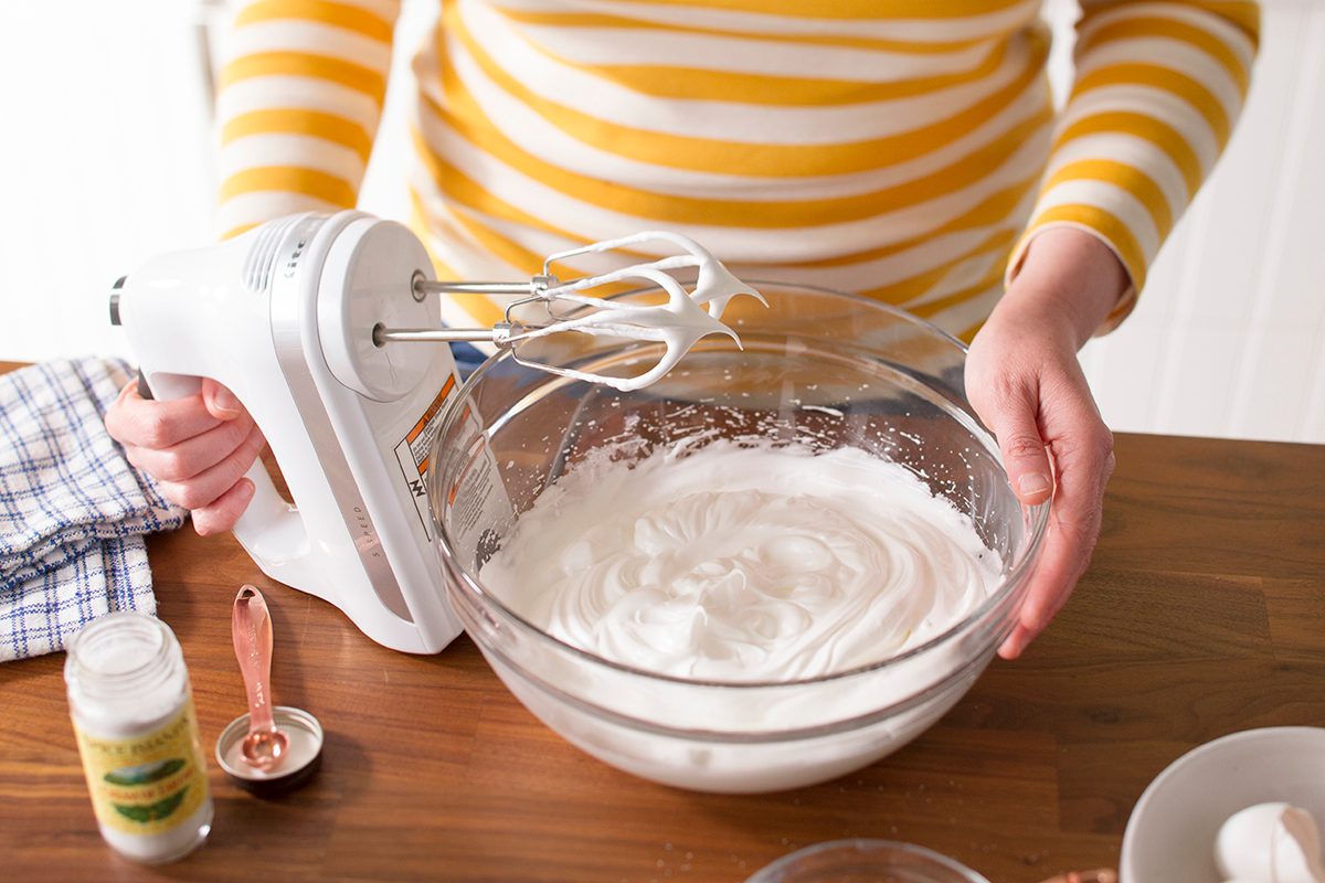A person in a yellow and white striped shirt uses a hand mixer to whip cream in a glass bowl on a wooden countertop