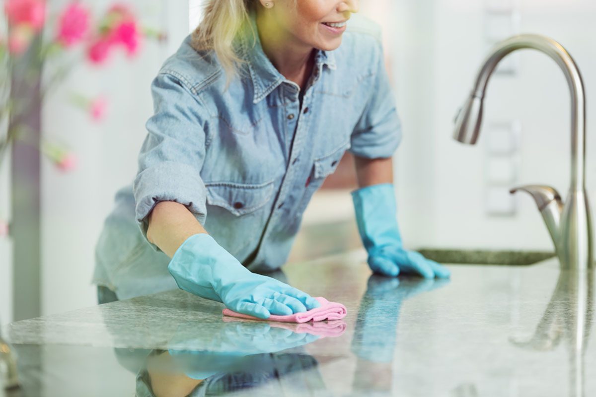 Woman wiping down kitchen countertop with pink cloth, wearing blue jean shirt and rubber gloves for household cleaning