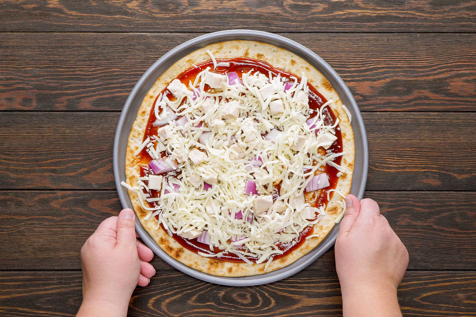 Someone Holding a Pizza Pan filled with Barbecue Chicken Pizza with hands on Wooden Surface