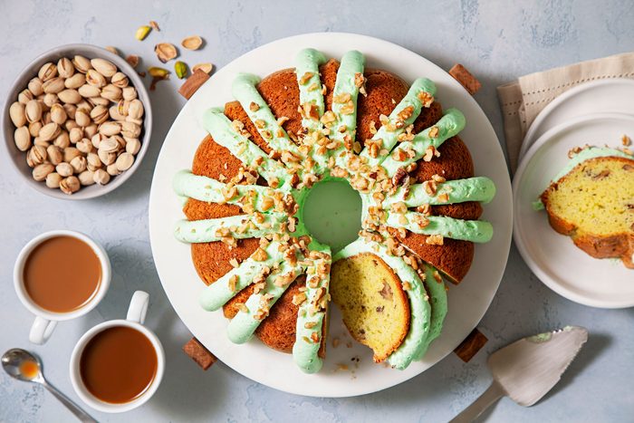 Pistachio Pudding Cake On A White Plate with A Bowl of Pistachio and Coffee Cups Next to it