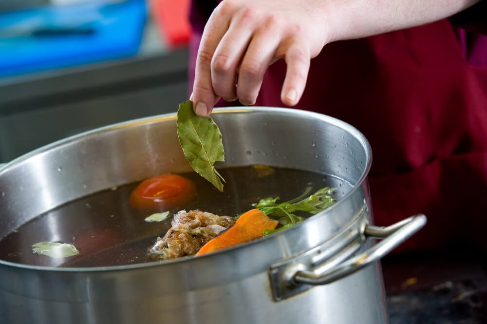 closeup hand of chef adding an aromatic bay leaf into pot with boiling vegetables