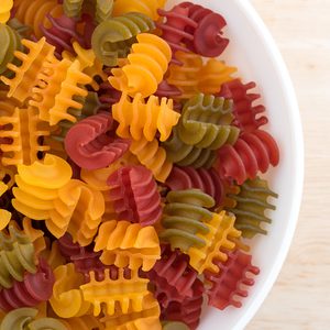 Top close view of a portion of gluten free corn vegetable radiatore pasta in a white bowl on a wood table top illuminated with natural light.