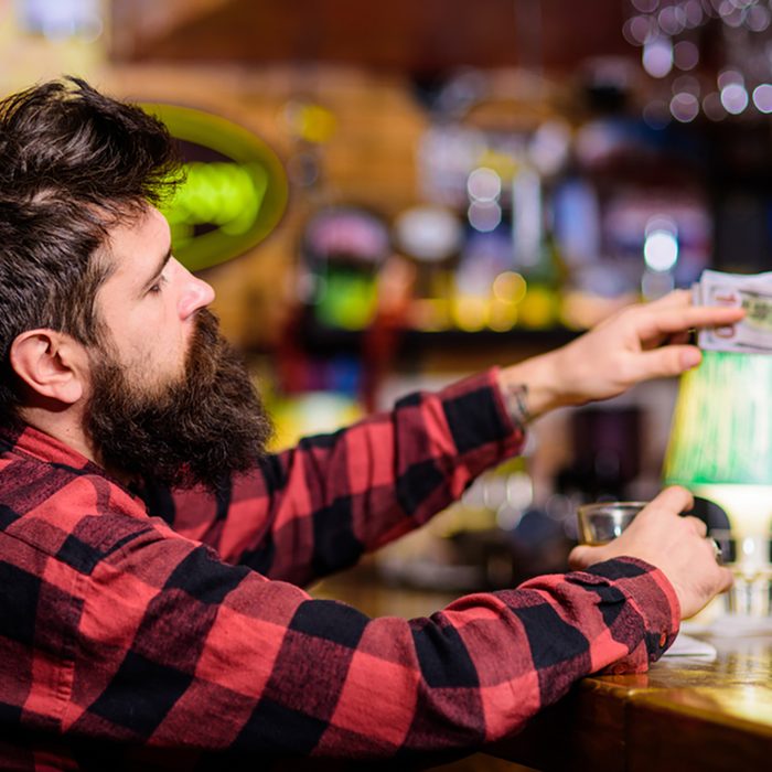Man with drunk face sit alone at bar counter. Hipster holds glass with alcoholic drink and money, ordering more drinks.