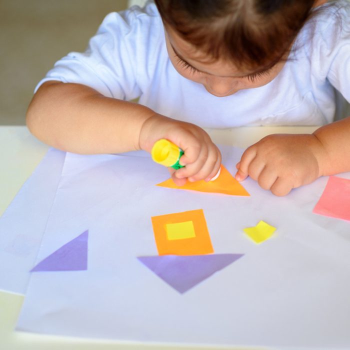 Cute little girl make applique, glues colorful house, applying a color paper using glue stick while doing arts and crafts in preschool or home.