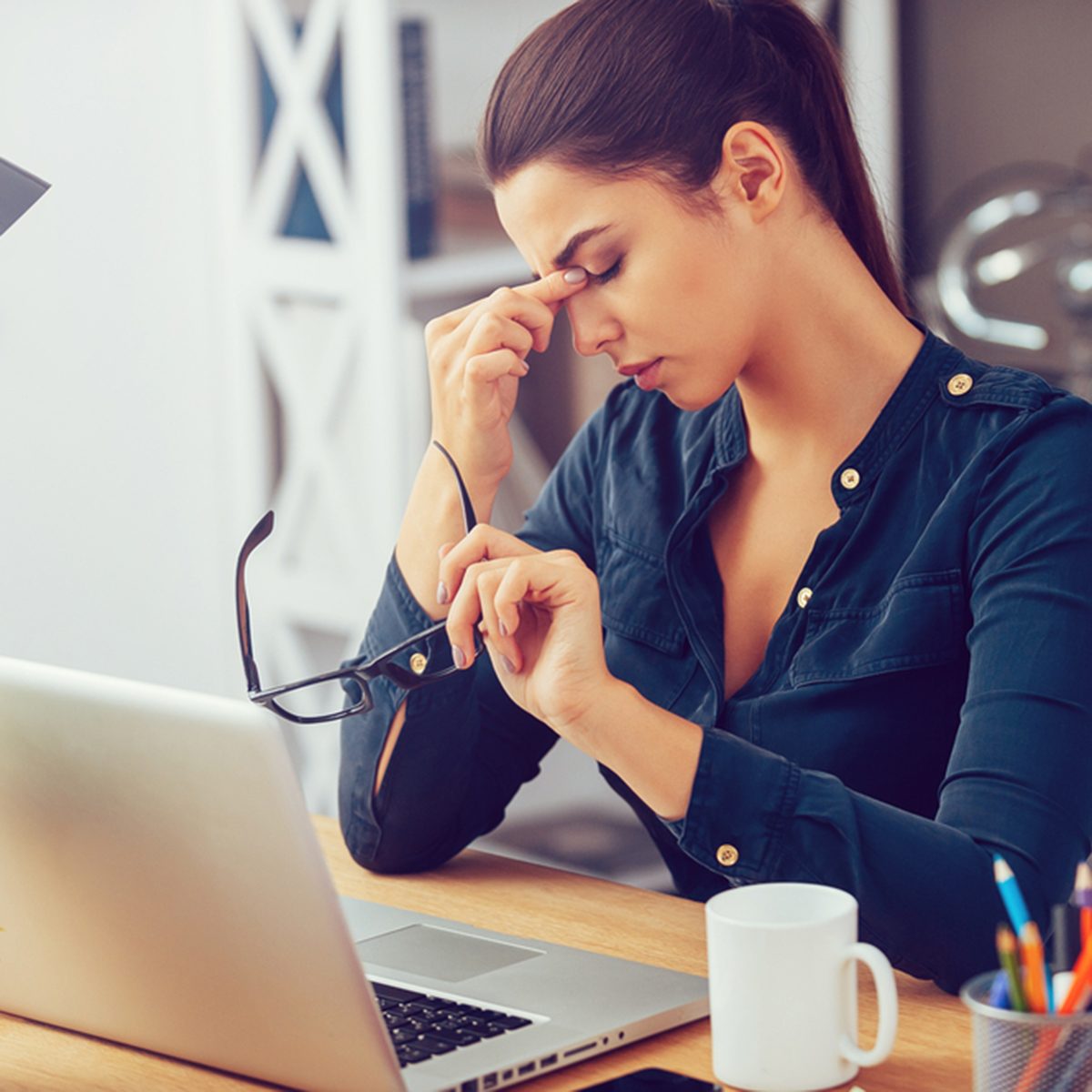Frustrated young woman keeping eyes closed and massaging nose while sitting at her working place in office