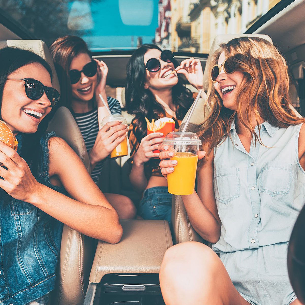 Enjoying their lunch in the car. Four beautiful young cheerful women looking at each other with smile and eating take out food while sitting in car