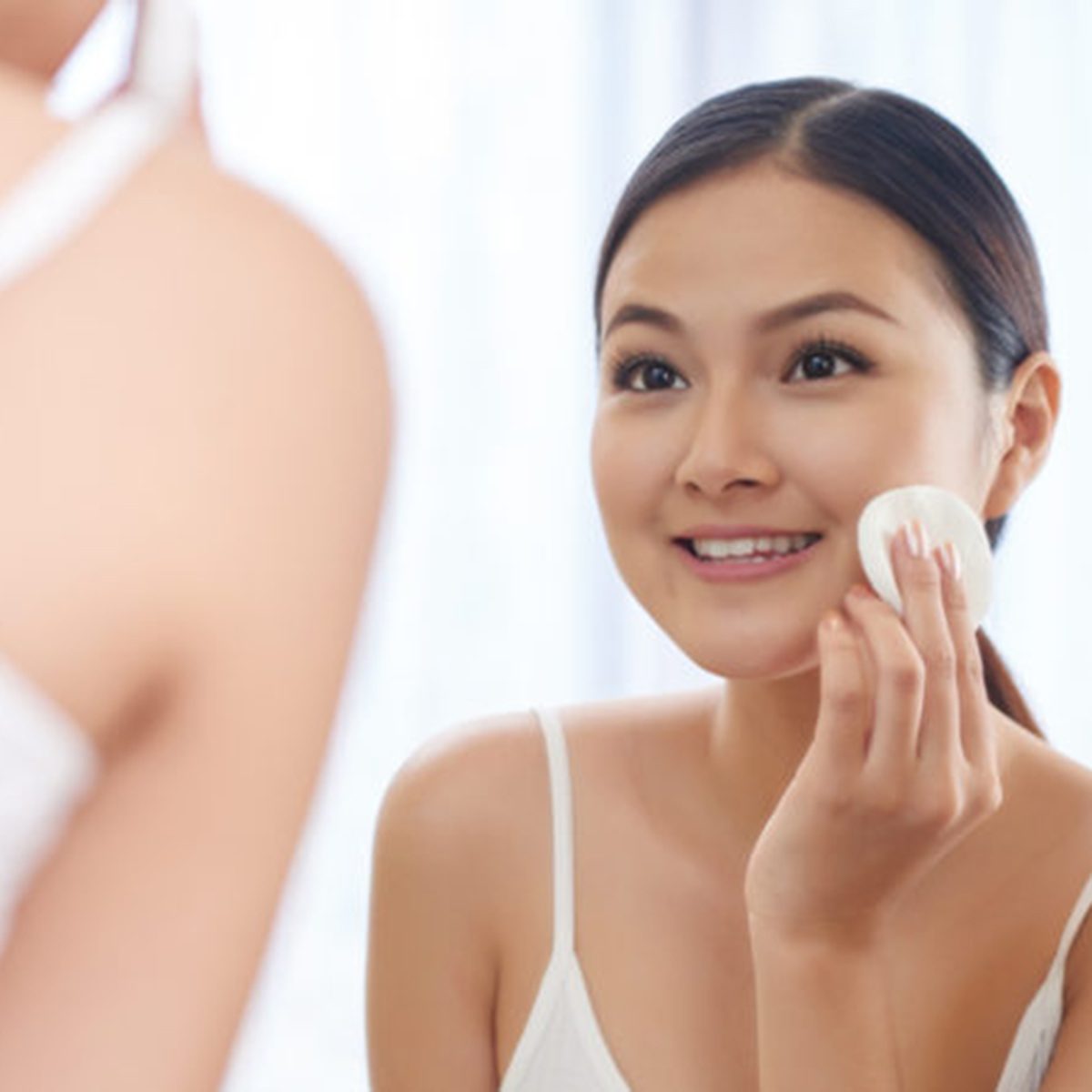 Smiling young woman applying toner on her face in front of mirror
