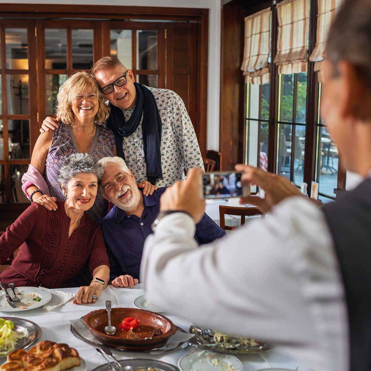 Group of senior friends meet up. They are in the restaurant, having lunch and taking. Waiter photographing with smart phone.