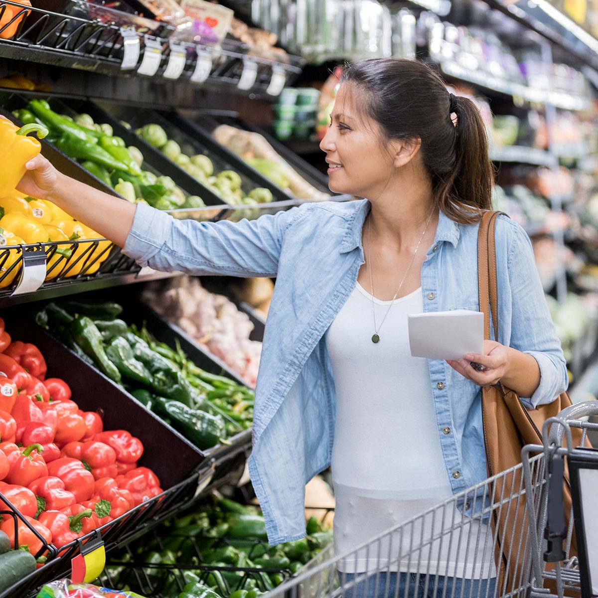 A smiling mid adult woman holds a grocery list as she shops for produce. She reaches to pick up a yellow pepper.