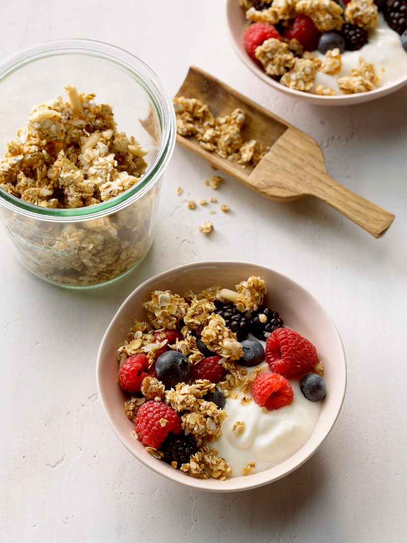 Easy Granola Recipe; high camera angle; white slight textured russian surface; raspberries; blueberries; Weck glass jar; small wooden scoop; Greek Yogurt