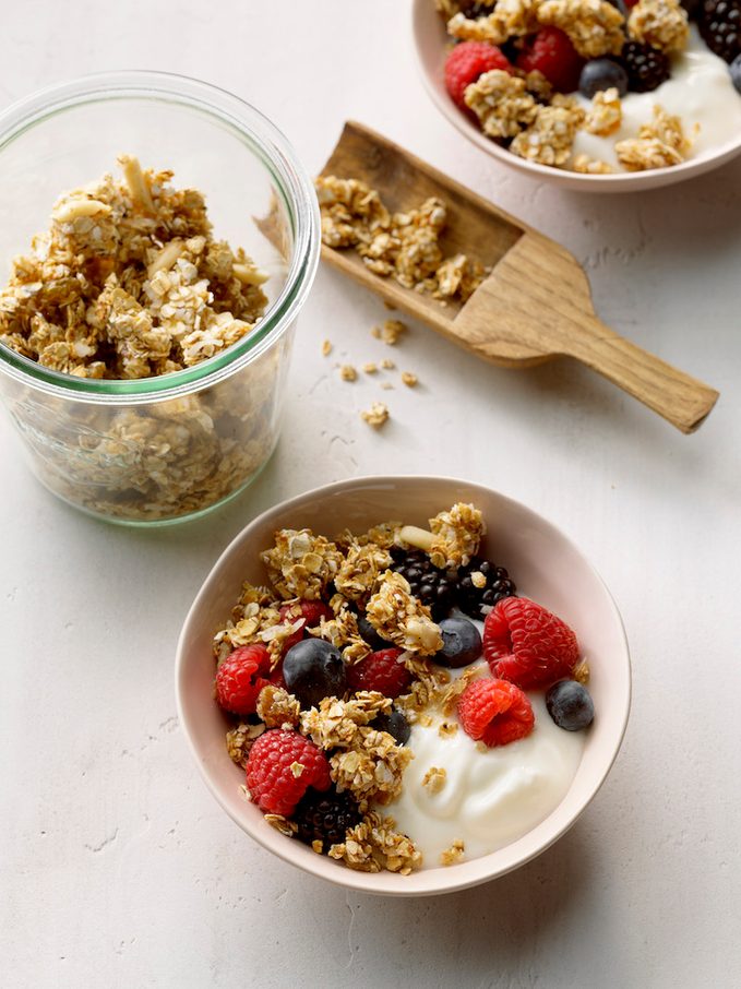 Easy Granola Recipe; high camera angle; white slight textured russian surface; raspberries; blueberries; Weck glass jar; small wooden scoop; Greek Yogurt
