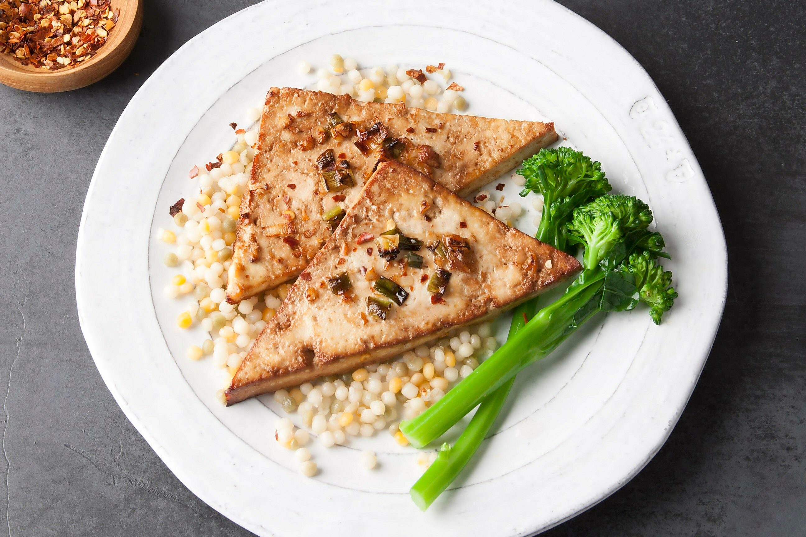 Three grilled tofu triangles served on a bed of pearl couscous with a side of steamed broccolini on a white plate. A small bowl of red pepper flakes is visible in the corner.