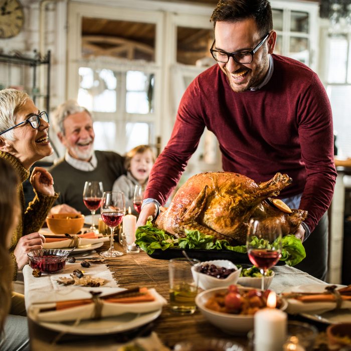 Happy man serving roasted turkey to his family during Thanksgiving dinner at dining table.