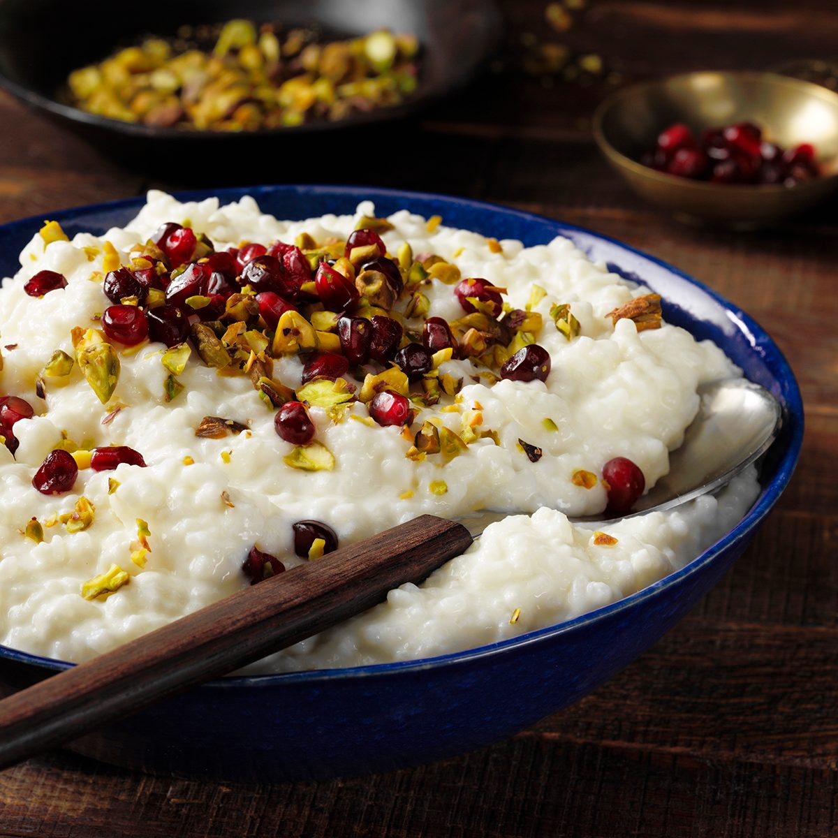 GLOBAL FLAVORS GO MAINSTREAM Food Trends Report, Rose Water Rice Pudding; 3/4 camera angle; wood surface; dark wood surface; russian wood surface; blue bowl; rose water; dark bowls; long grain rice; half-and-half cream; rose water; Pomegranate seeds; chopped pistachios; wood handled spoon; spoon; serving spoon