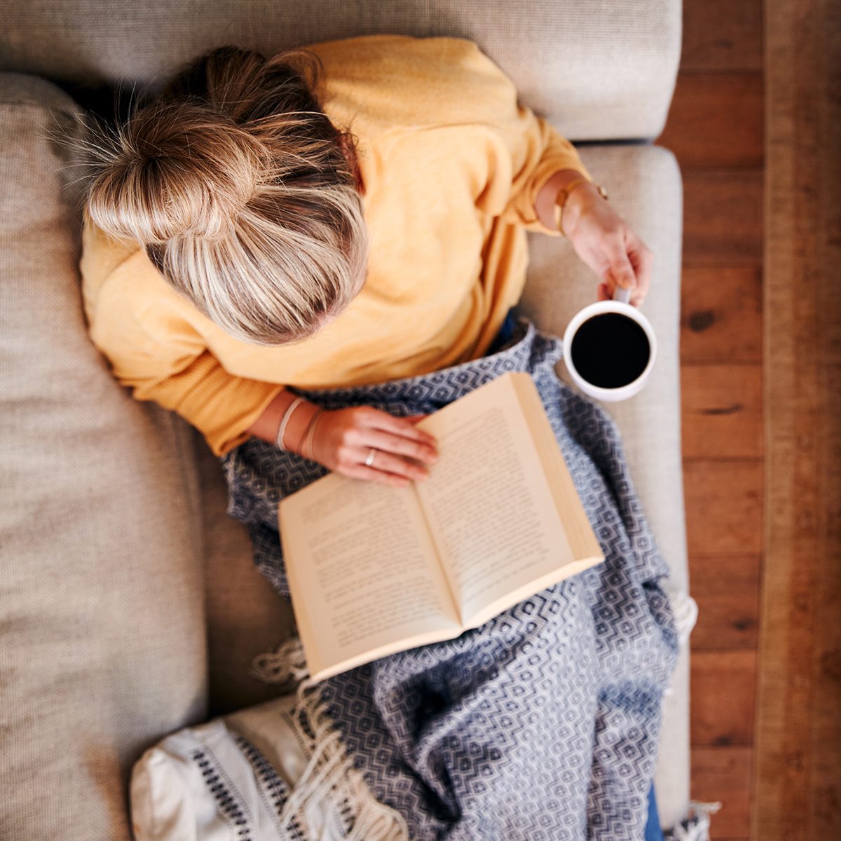 Overhead Shot Looking Down On Woman At Home Lying On Reading Book And Drinking Coffee