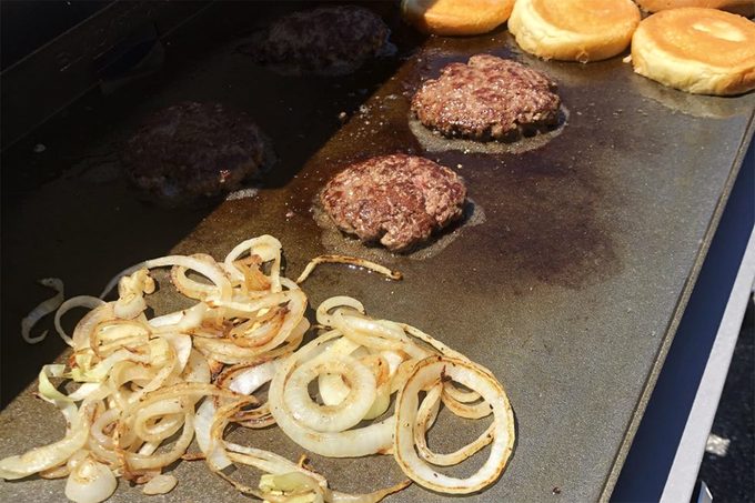 Sliced onions, two hamburger patties, and toasted buns cooking on a flat-top grill.
