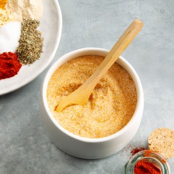 Curry Powder Seasoning Mix in a white bowl with a wooden spoon; in the corner is a partial view of a plate with different colored seasonings on it