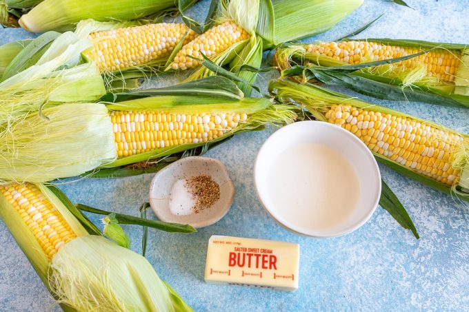 corn on the cob, butter, a dish of spices and a bowl of milk on a blue countertop