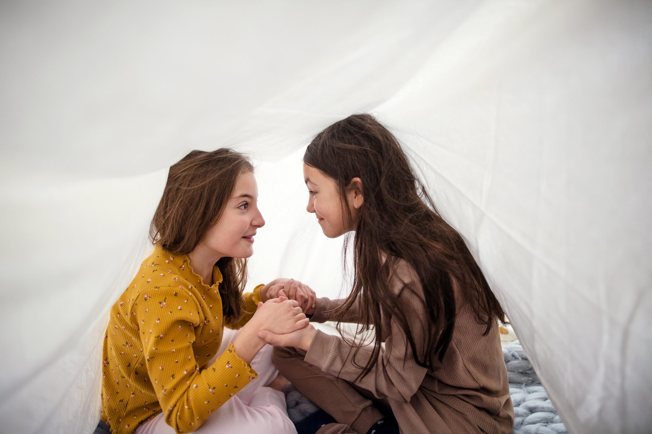Two small girl friends sitting under duvet on bed, holding hands and talking.