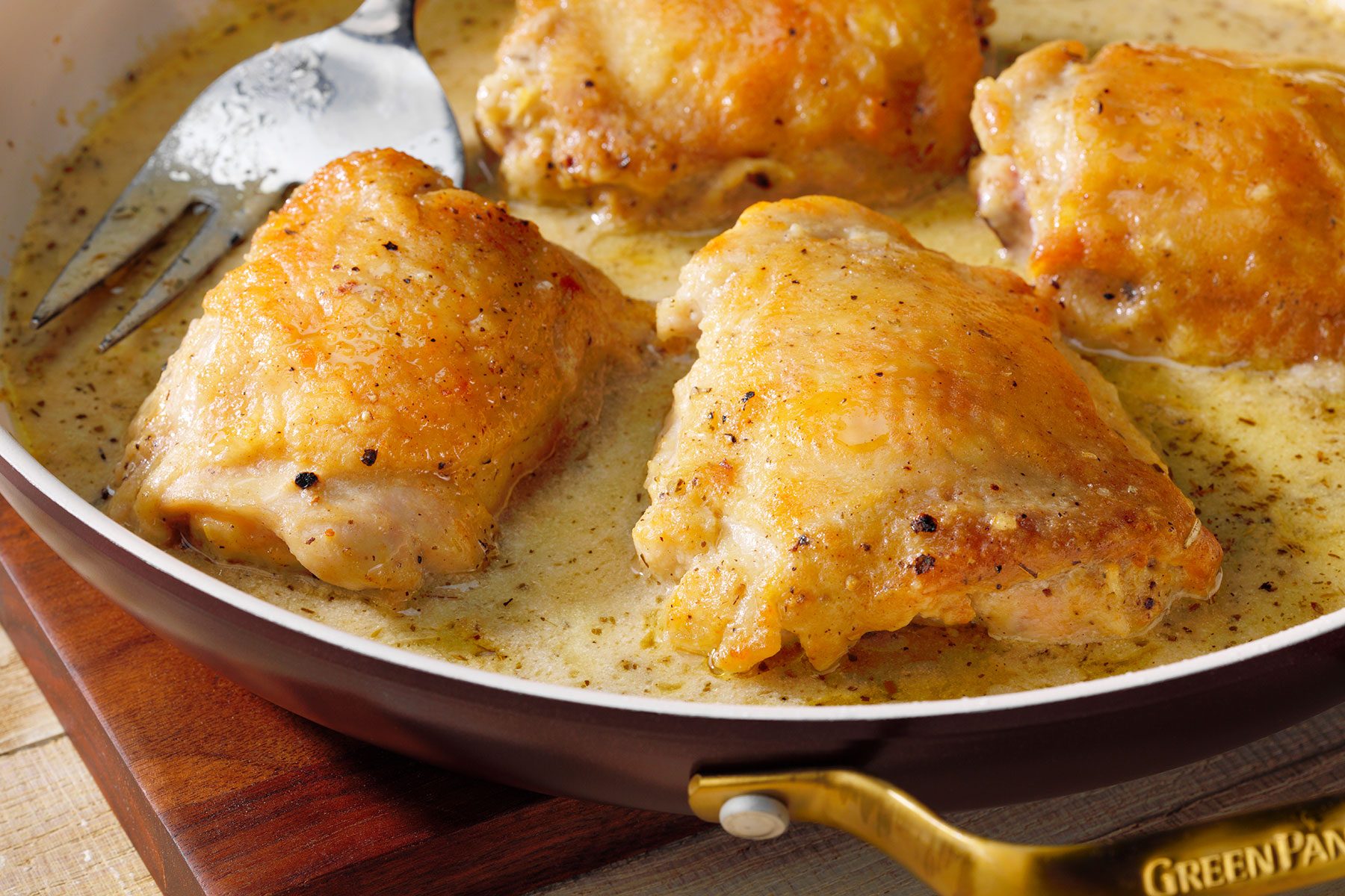 A sauté pan containing four pieces of browned, seasoned chicken thighs bathed in a creamy sauce. A silver fork rests in the sauce next to one of the chicken pieces. The pan handles are gold-colored and the pan sits on a wooden board.