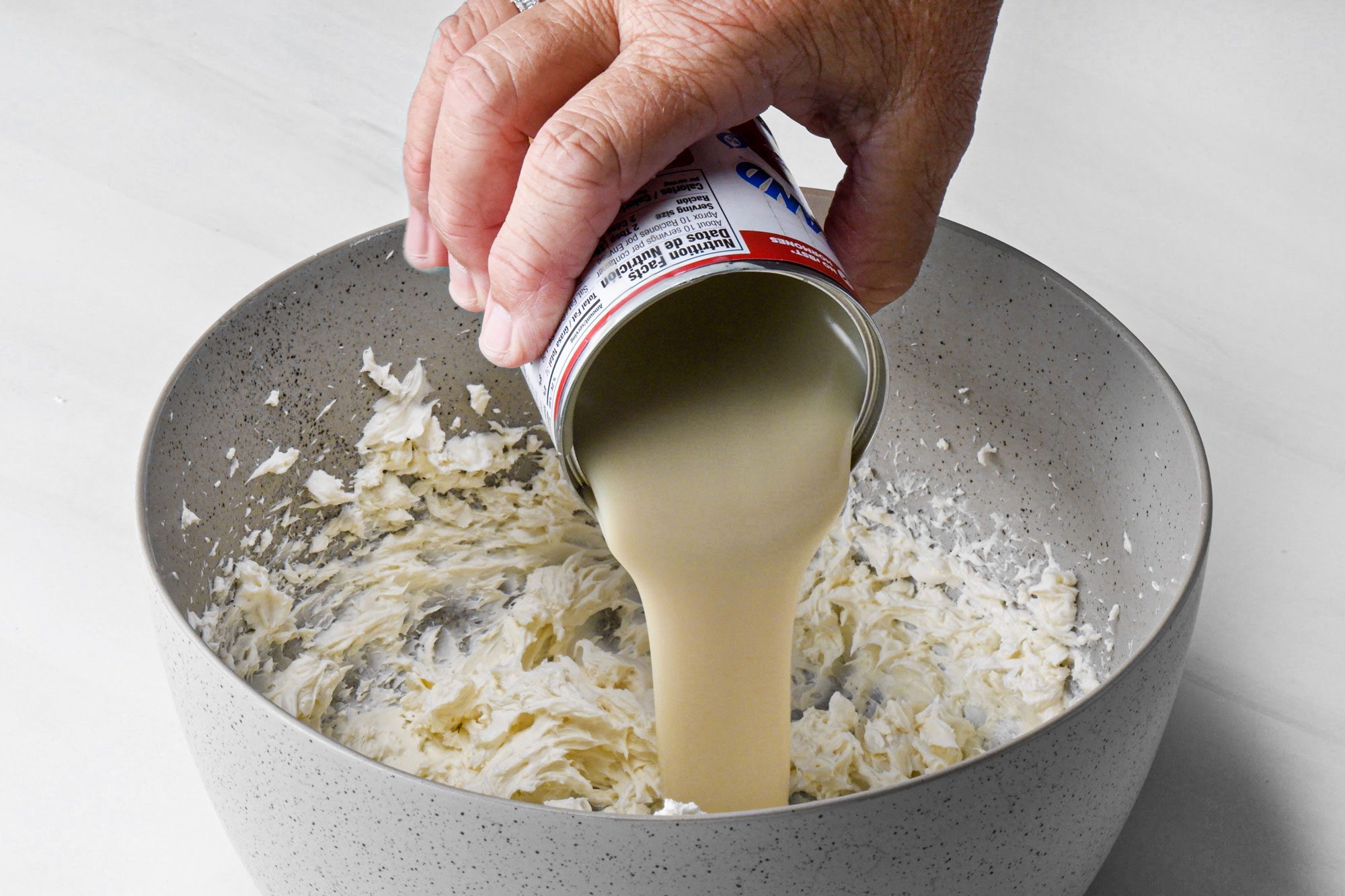 a person pouring sweetened condensed milk in large bowl on marble surface