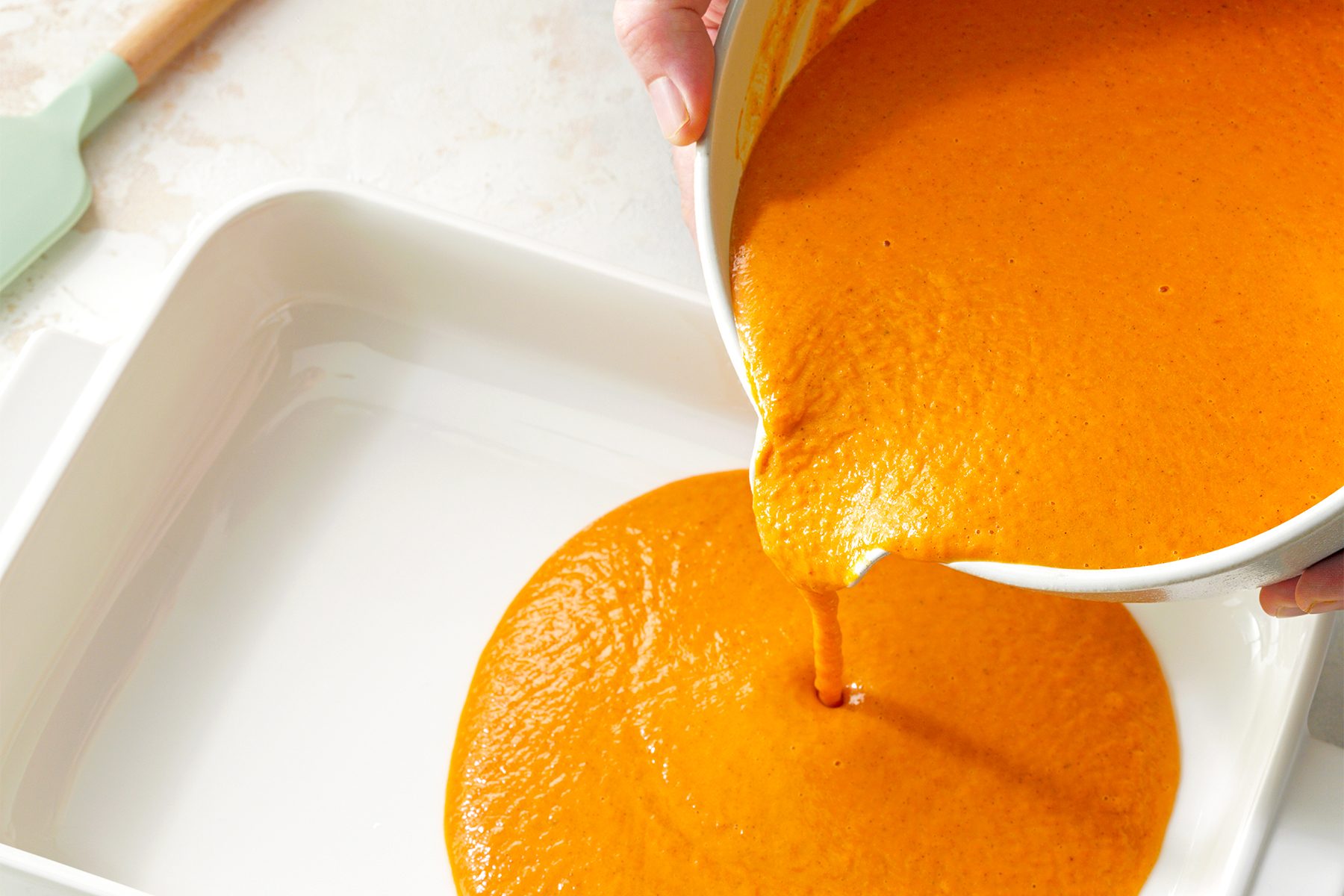 A person pours a smooth, orange batter from a white bowl into a white rectangular baking dish