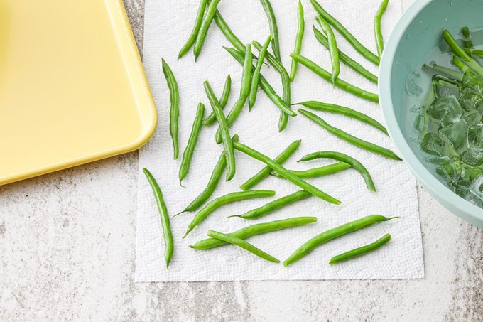 Overhead view of cooled green beans being spread on a paper towel to dry and continue prepping for freezing. Baking tray nearby for the next step.