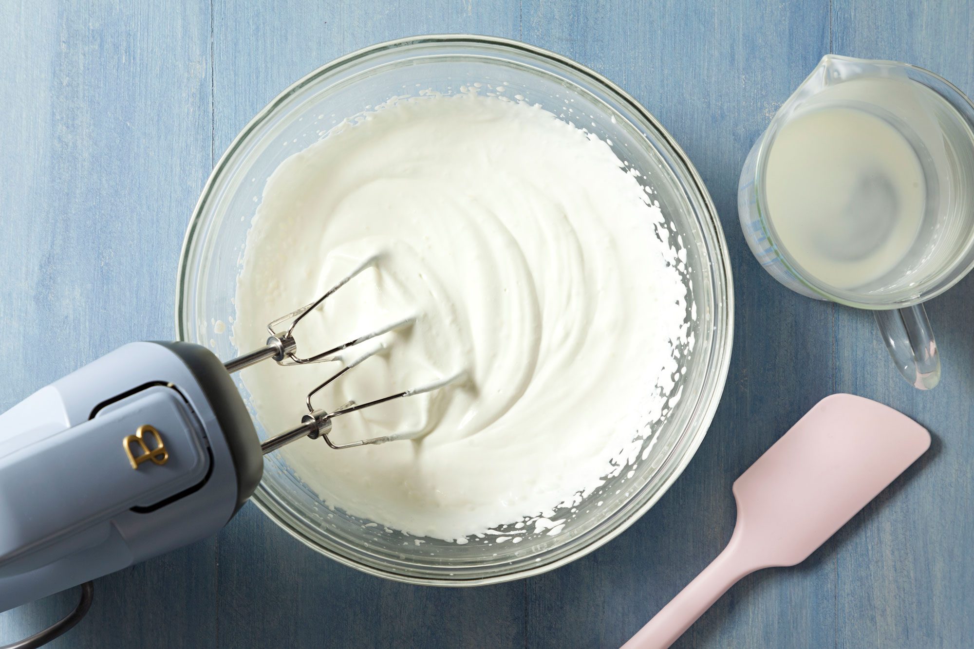 beating cream In a large glass bowl using a hand mixer on a textured light blue surface