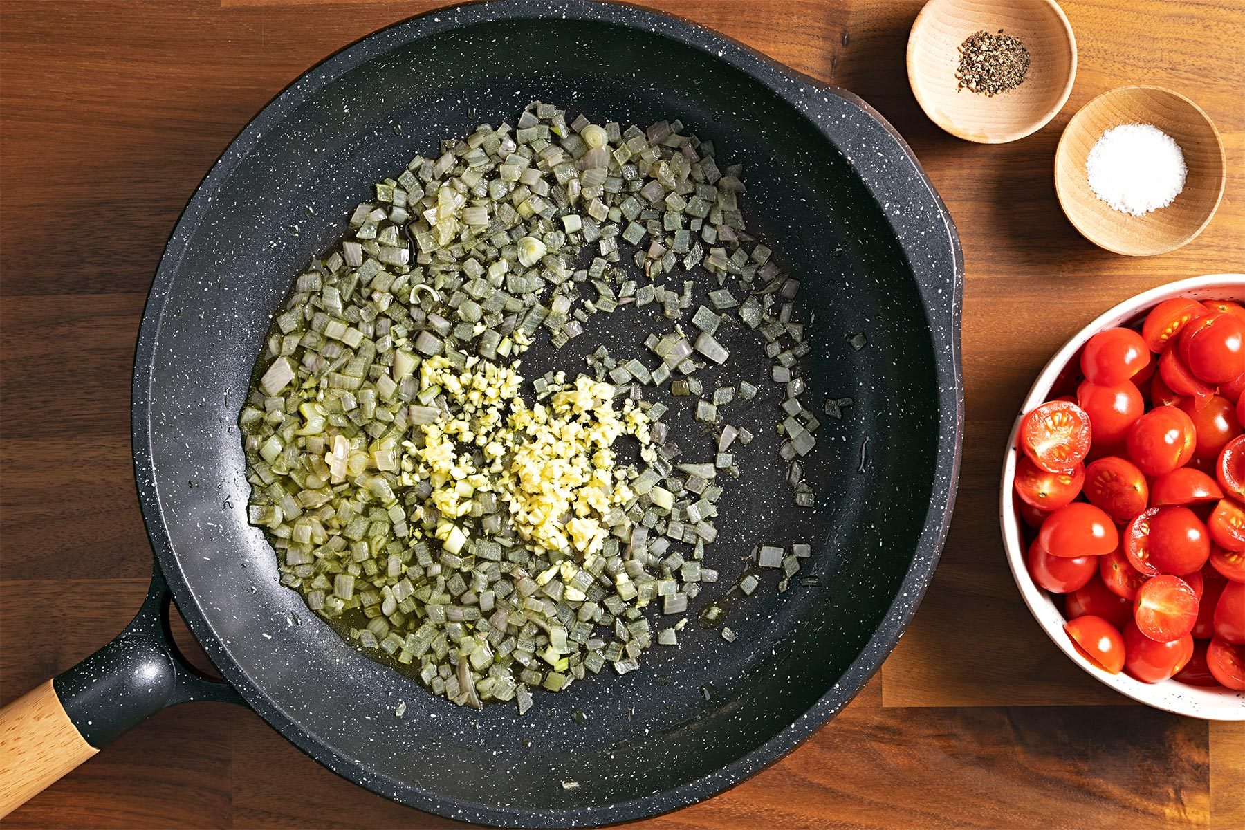 Overhead shot of preparation for topping; large skilletr; oil; garlic; tomatoes bowl; a small bowl of salt and pepper; brown wooden background;