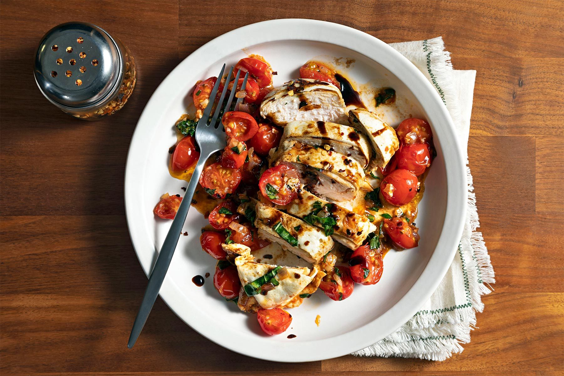 Overhead shot of Baked Bruschetta Chicken; served on plate; serve prepared topping over chicken; sprinkle with red pepper flakes and chopped basil; fork; chilli flakes bottle; napkin; brown wooden background;