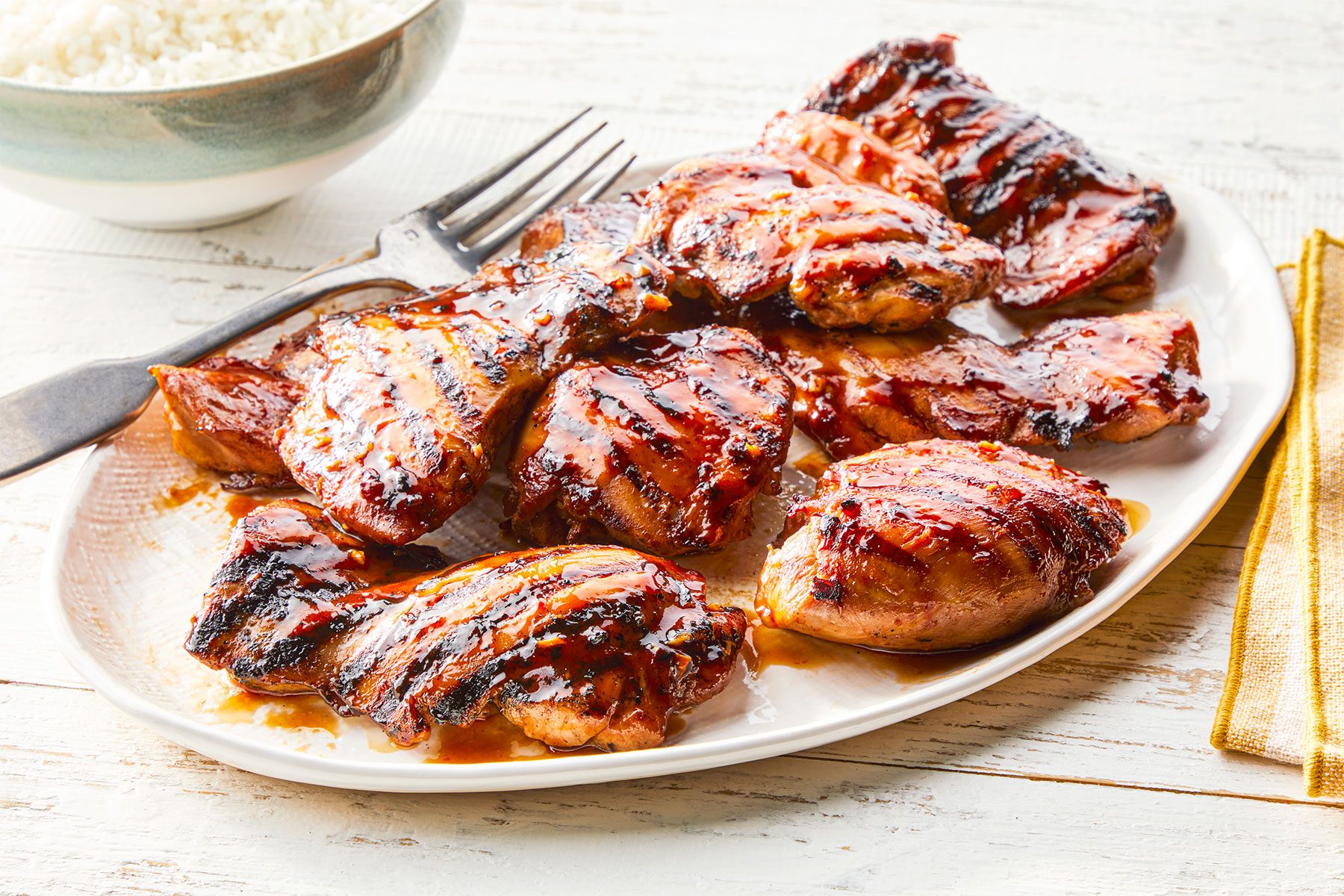 Overhead shot of Grilled Huli Huli Chicken; served on large platter; fork; rice bowl; napkin; white wooden background;