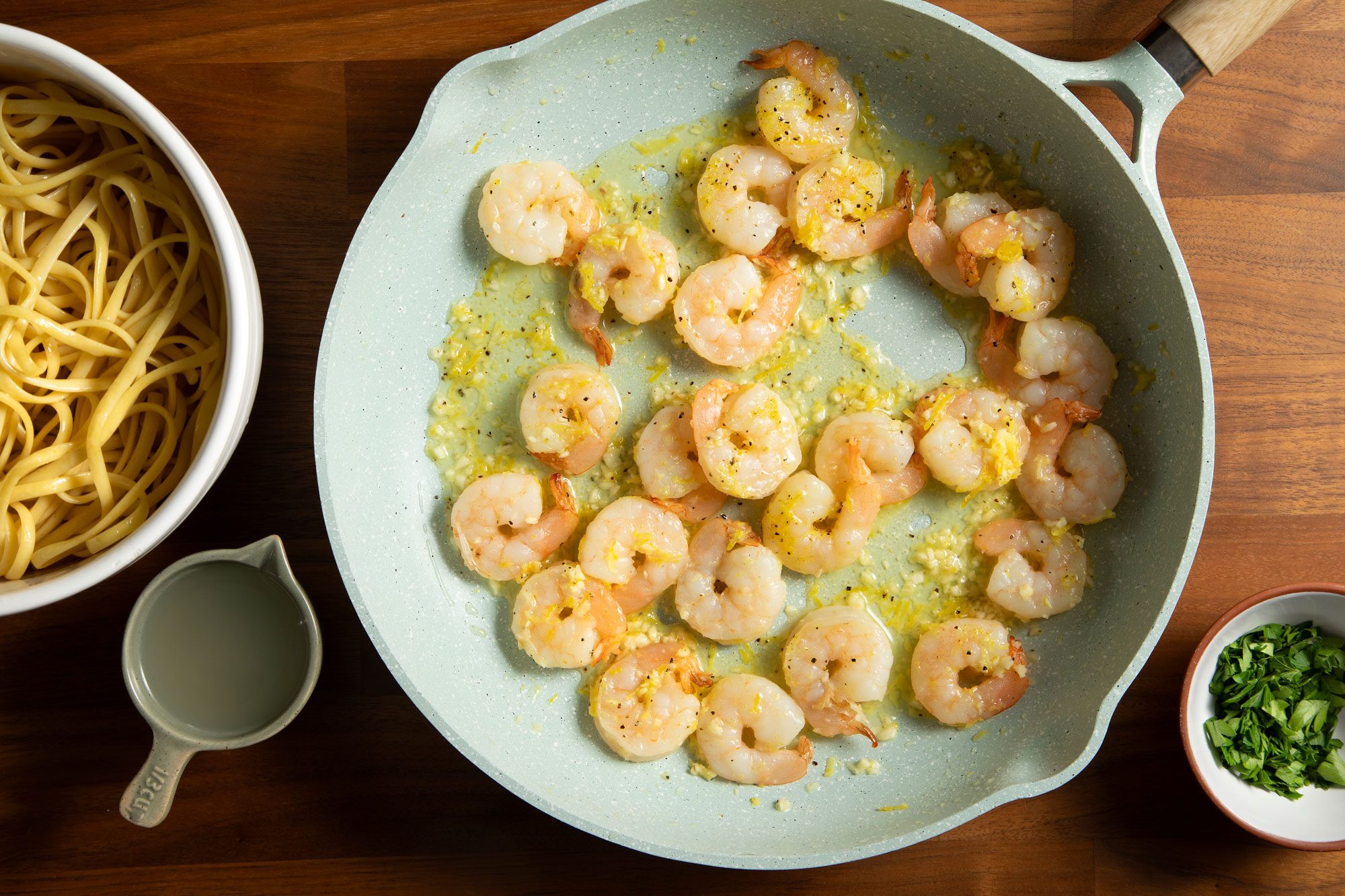 overhead shot; wooden background; cooking shrimps in large white pan with some garlic; pasta in a bowl;