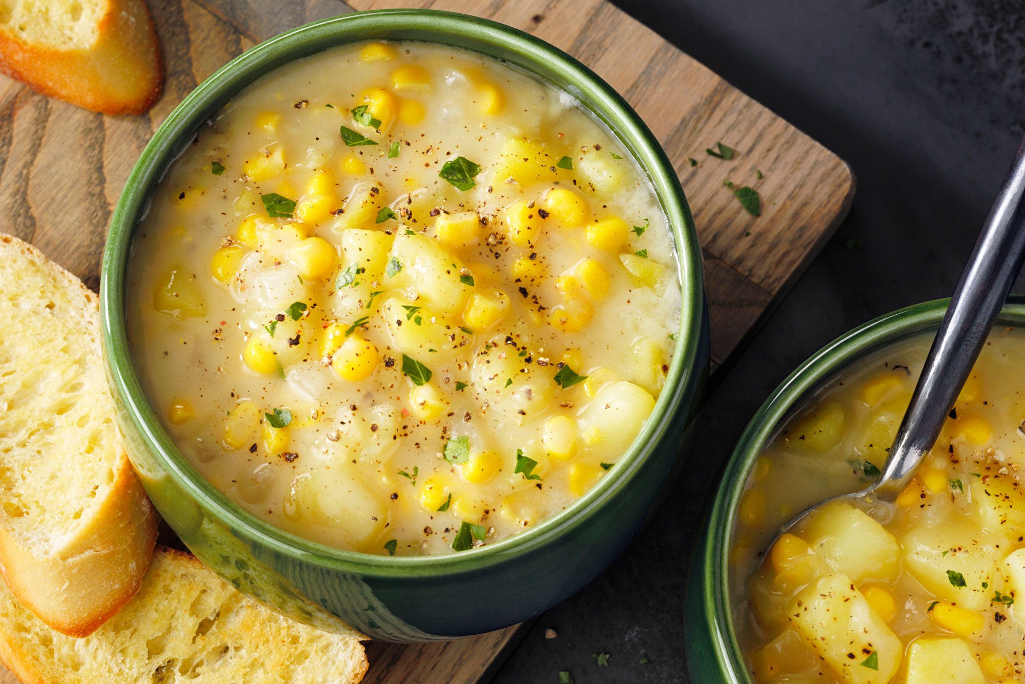 Two bowls of Quick Potato Corn Chowder with bread on a wooden cutting board topped with chilli flakes and minced parsley