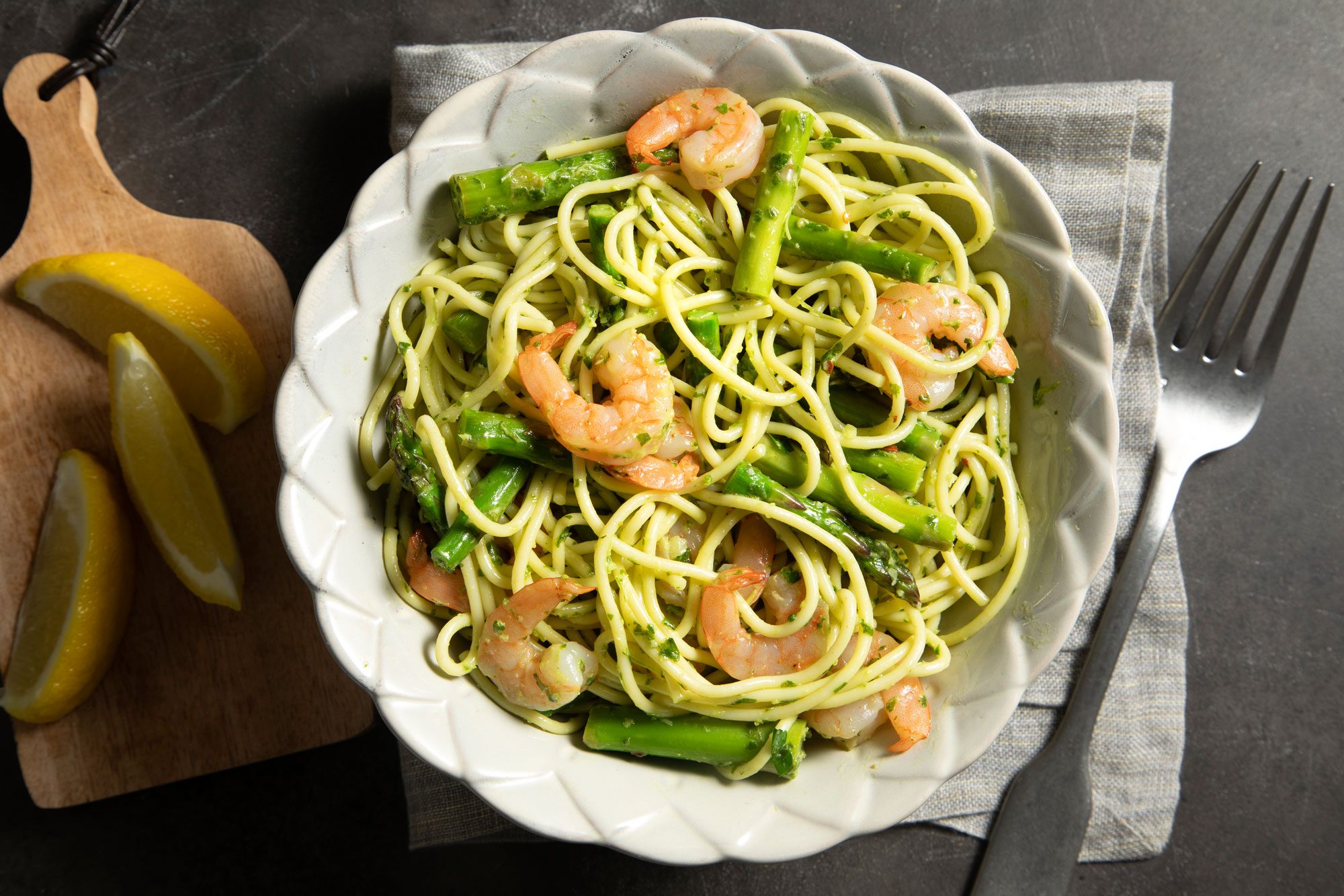 overhead shot; black background; Pesto Shrimp Pasta served on white plate with silver fork; lemon wedges on wooden board; over kitchen towel;