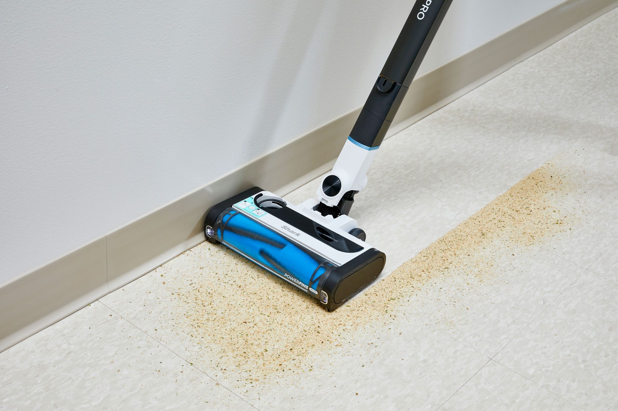 A floor-cleaning device with a blue and white body is vacuuming up light brown debris from a beige tiled floor near a wall. The appliance is positioned to clean precisely along the edge.