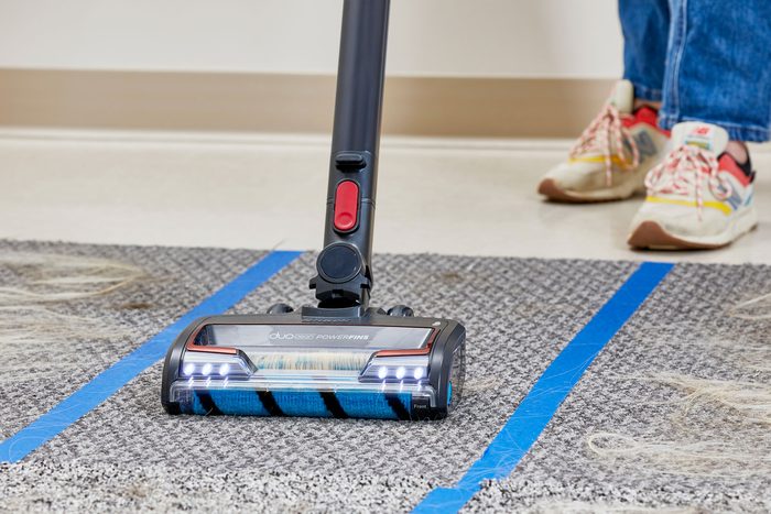 A person wearing sneakers is using an upright vacuum cleaner on a carpet with visible pet hair. The vacuum has LED lights and is positioned over two blue tape lines, showing the difference between cleaned and uncleaned areas.