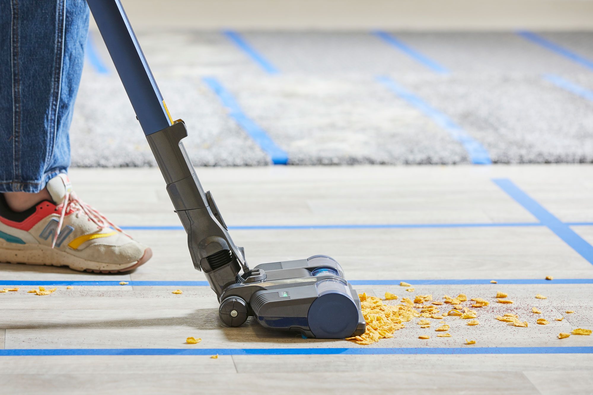 A person wearing blue jeans and sneakers uses a blue and gray vacuum cleaner to pick up scattered cereal pieces from a light-colored wooden floor. The floor is marked with blue tape lines, and part of a gray rug is visible in the background.