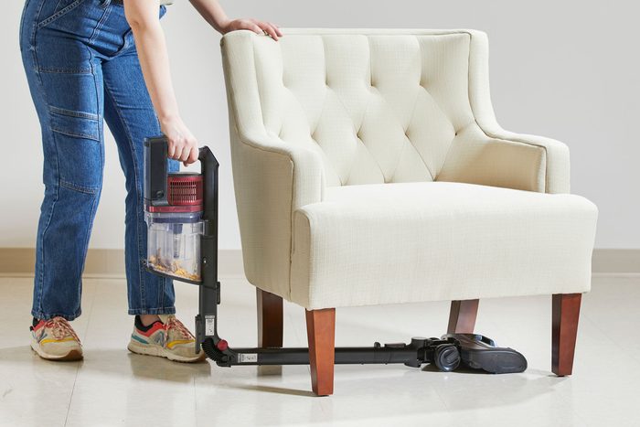 A person in jeans and sneakers uses a vacuum cleaner to clean the floor under a cream-colored, tufted armchair. The vacuum has a slim head, allowing it to reach easily beneath the chair.