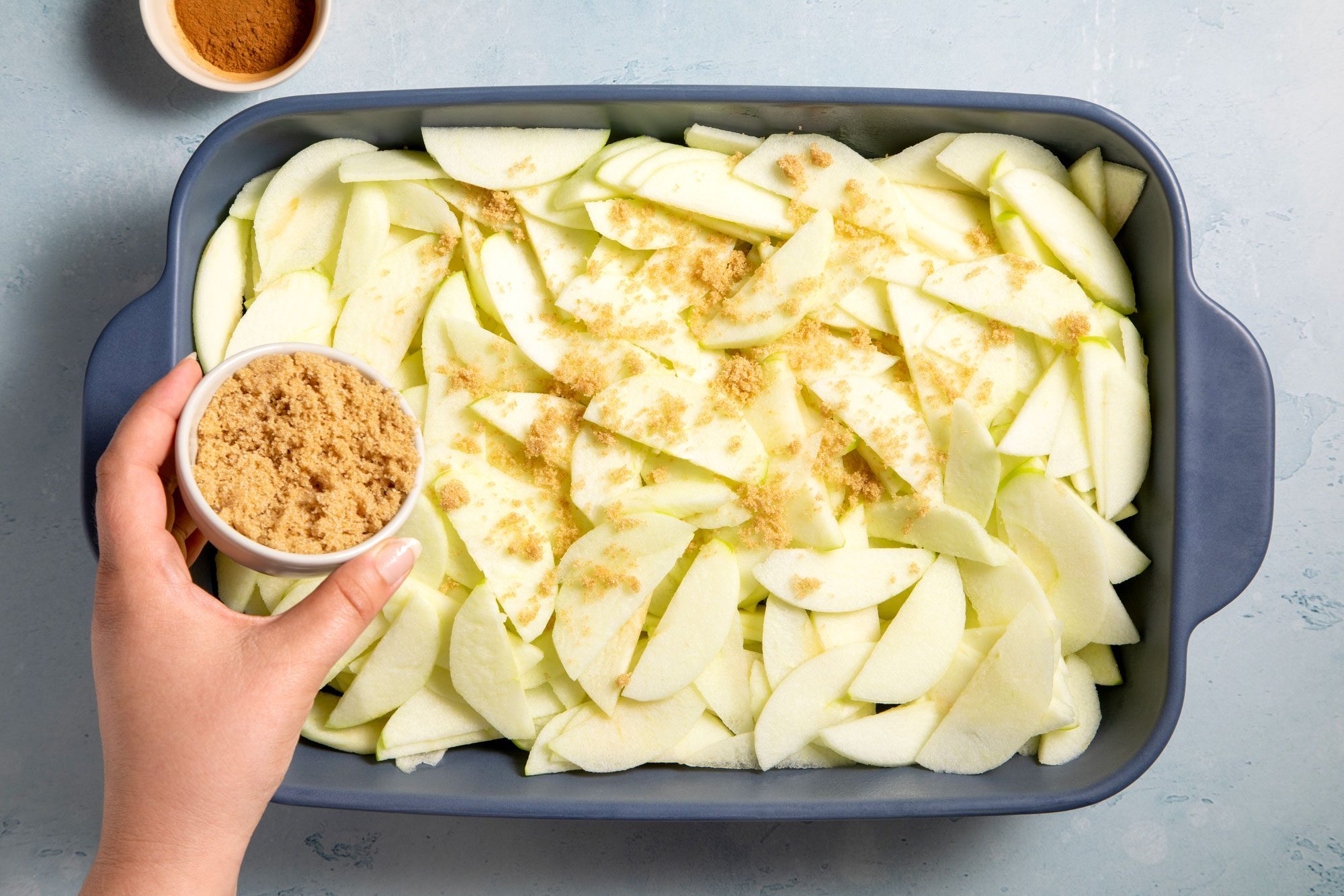 overhead shot; light blue textured background; Placed apples in a greased baking dish, Sprinkling brown sugar and cinnamon;