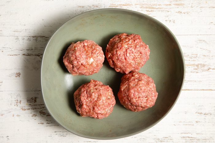 A green bowl contains four raw, seasoned meatballs placed on a white wooden surface.