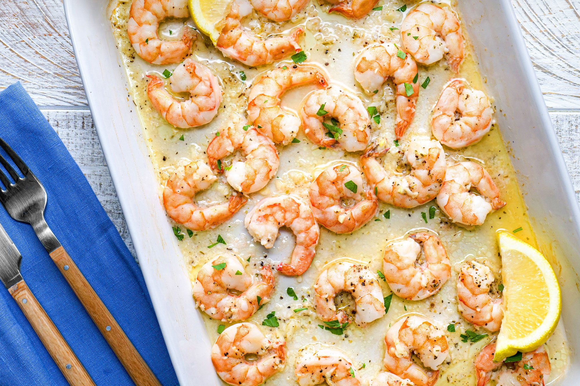 overhead shot; white wooden background; baked shrimps in white rectangular dish; with lemon wedges; two forks on blue napkin;