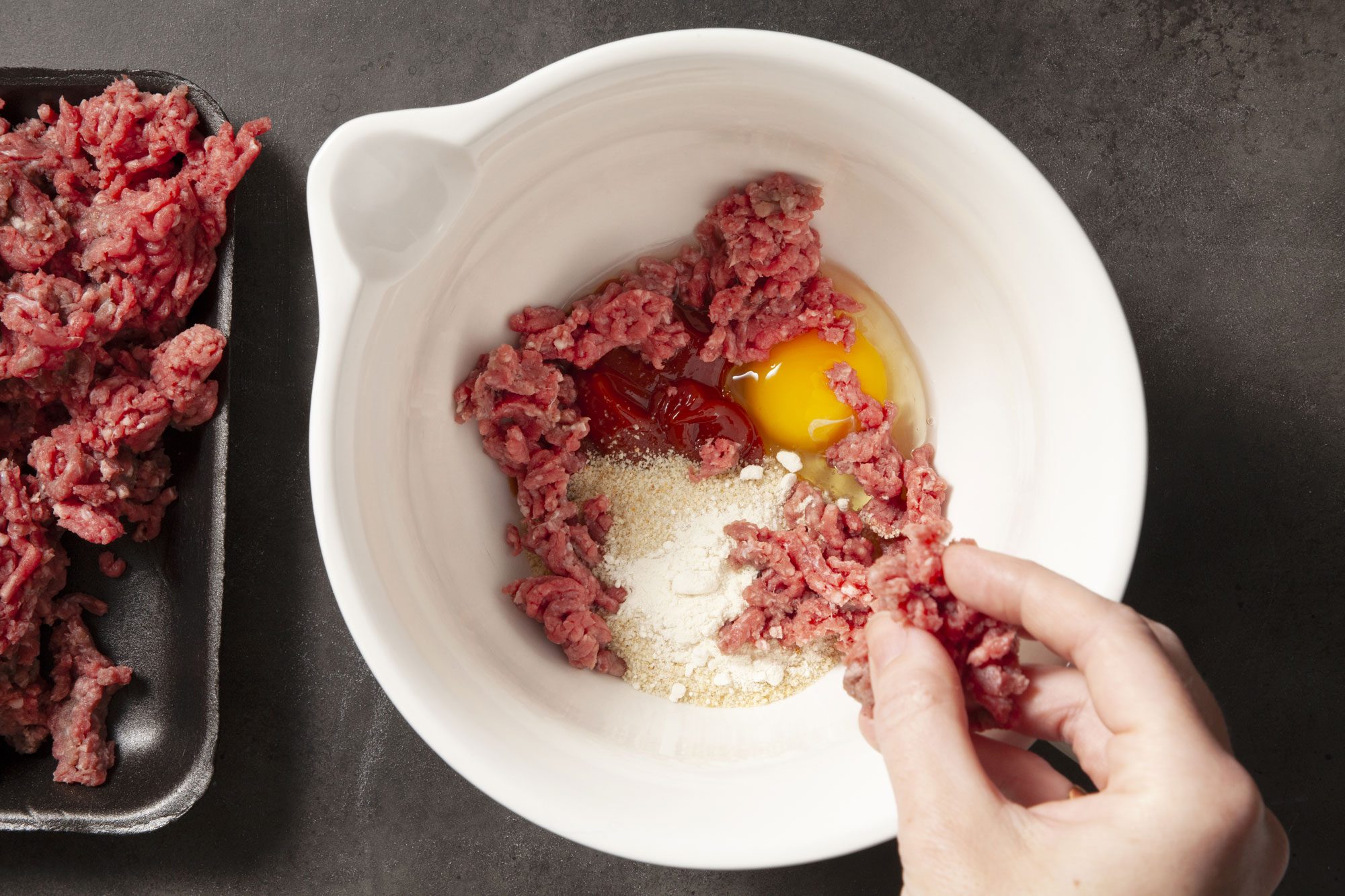 Overhead shot of combine 1/4 cup ketchup, egg, bread crumbs and onion powder in a bowl; crumble beef over mixture and mix well; black texture background;