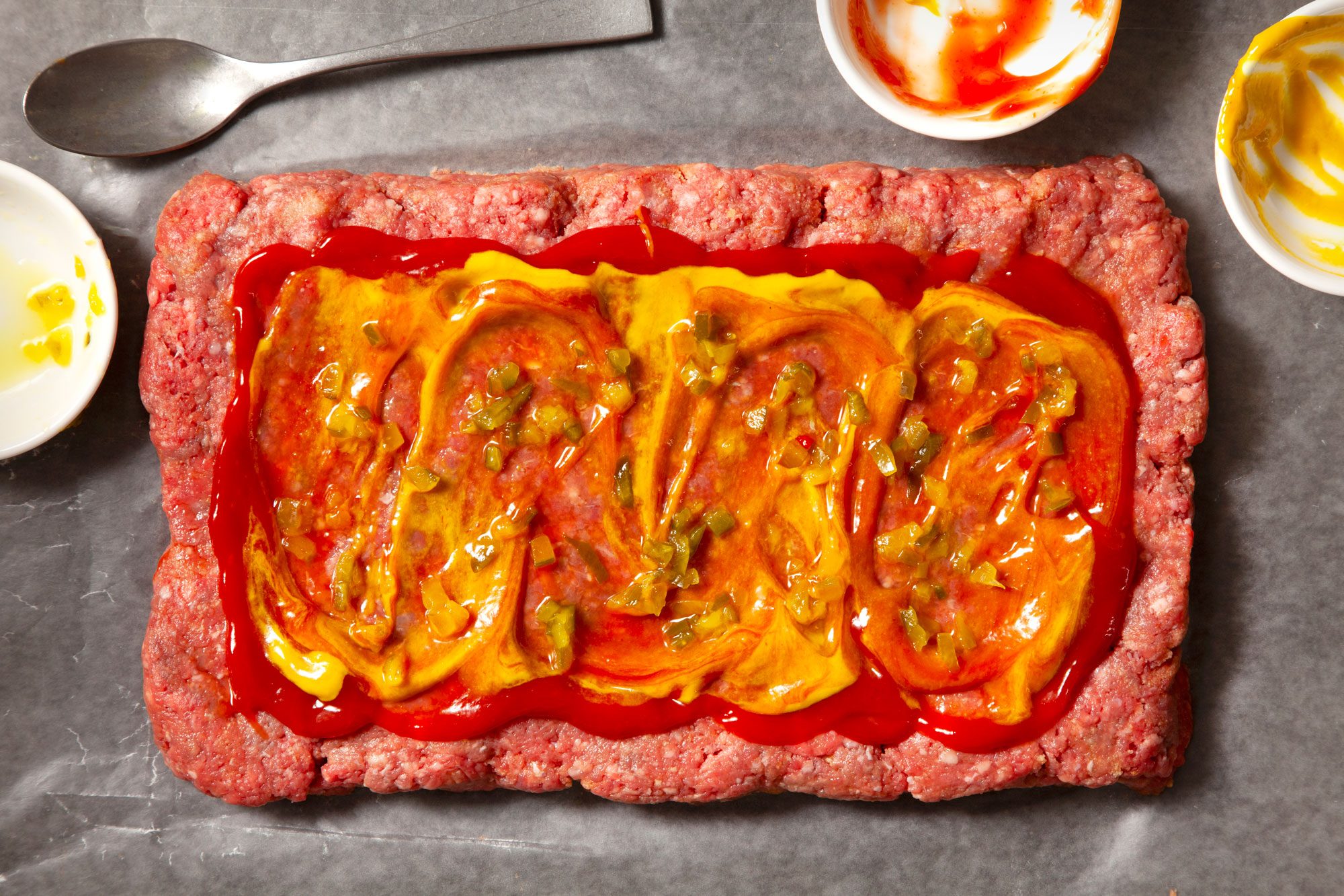 Overhead shot of a large piece of waxed paper; pat beef mixture into a rectangle; and spread remaining ketchup; top with mustard and relish; spoon; empty small bowls; black texture background;