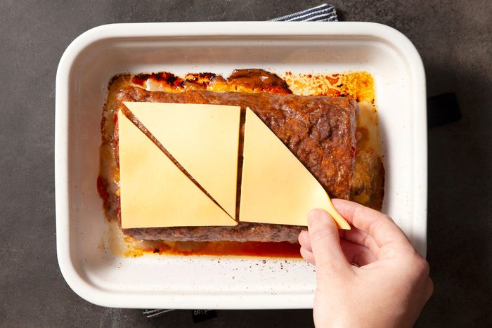 Overhead shot of cut the reserved cheese slices in half diagonally; place on top of loaf; baking pan; white and black line base cloth; black texture background;