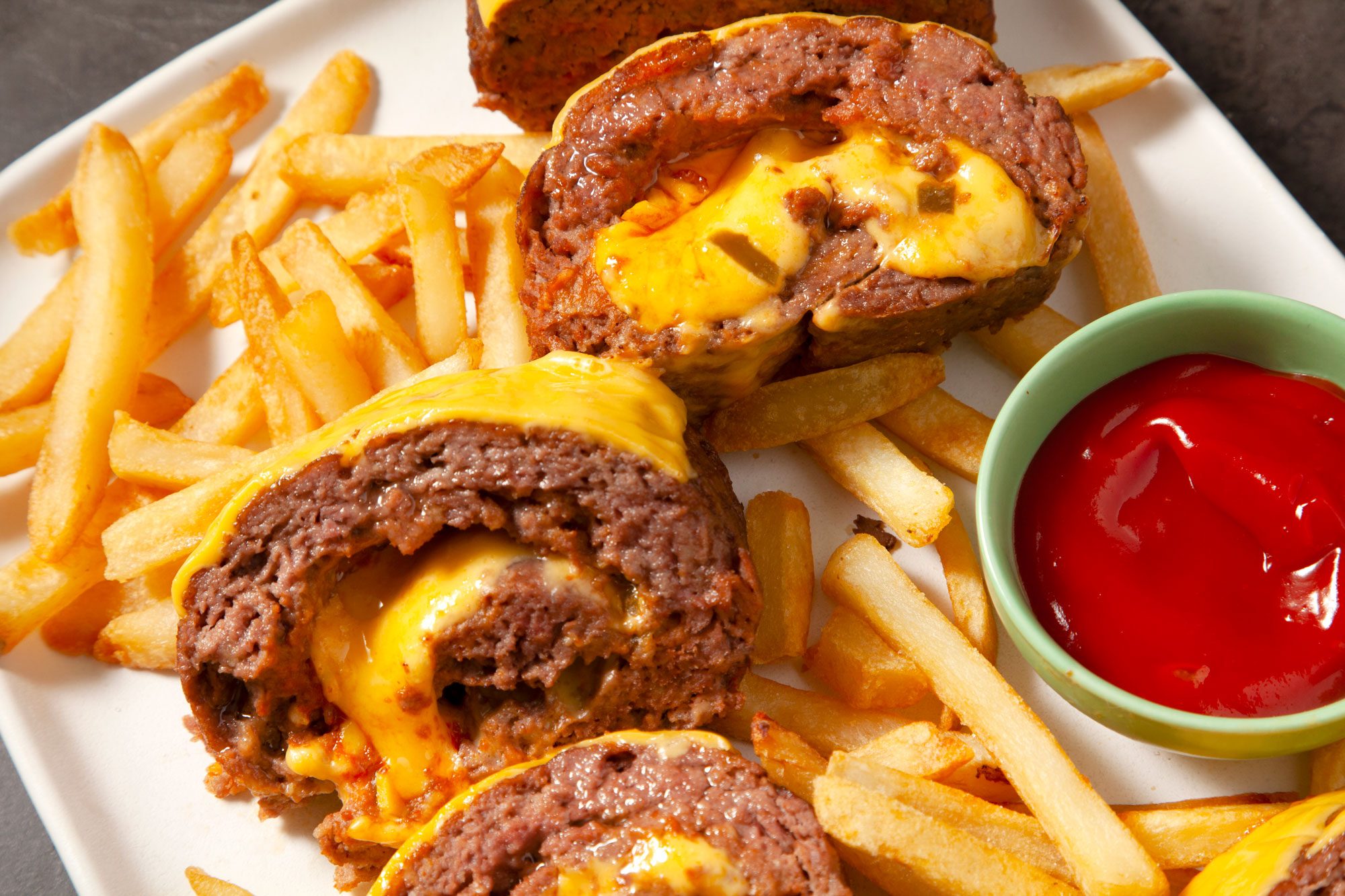 Close shot of Cheeseburger Meat Loaf; cut into small round pieces; served on large platter with fries and ketchup; black texture background;