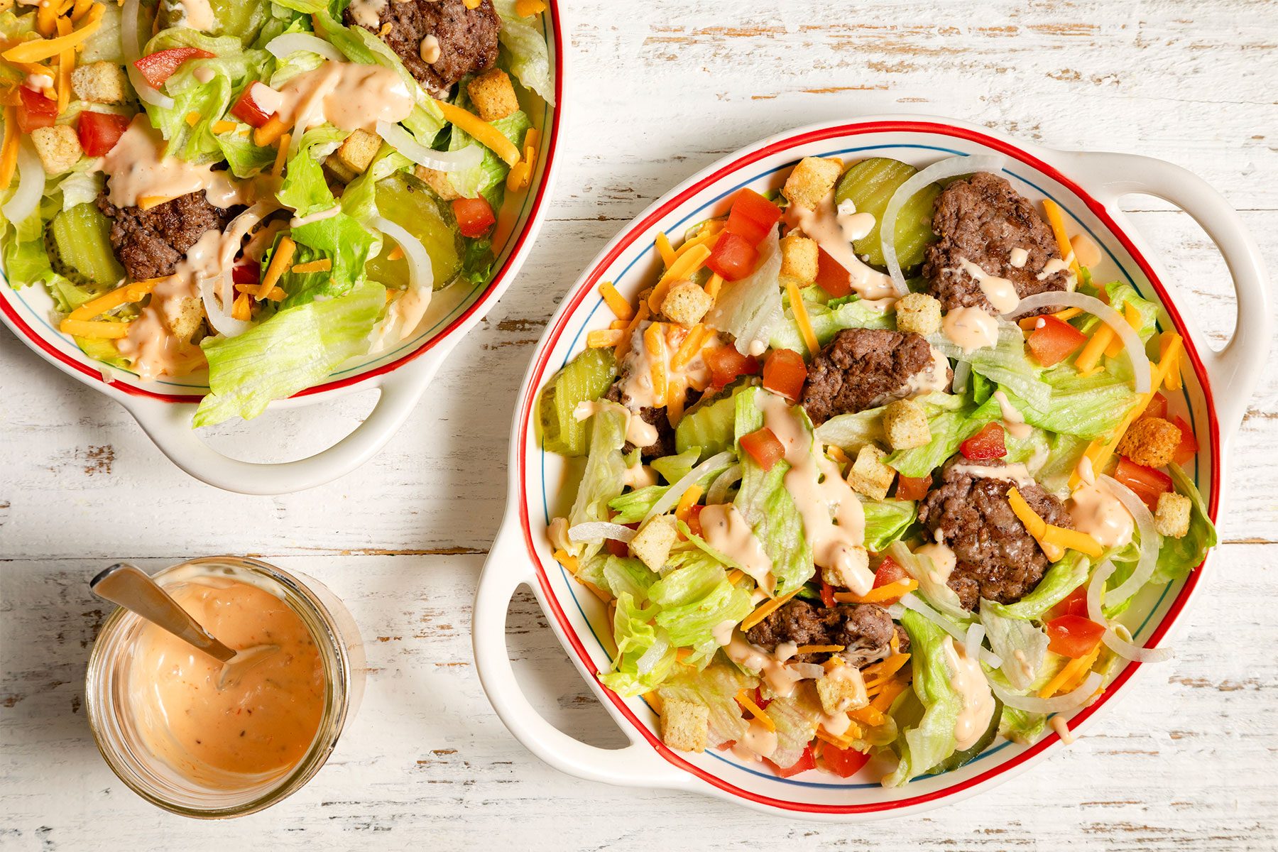 overhead shot; white textured background; 2 bowls of Deluxe Cheeseburger Salad with salad dressing;
