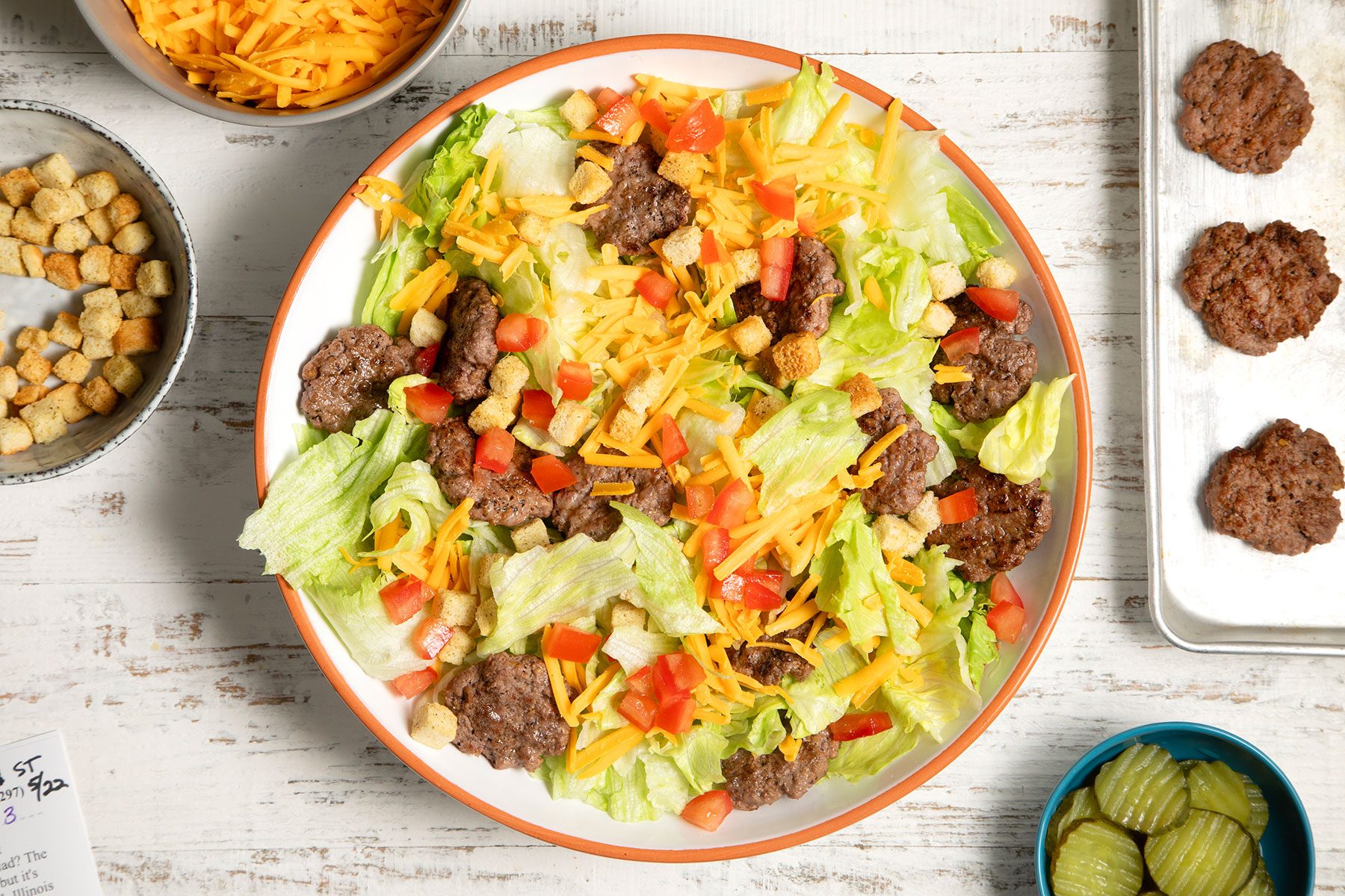 overhead shot; white textured background; Deluxe Cheeseburger Salad in a large bowl with fried patties, pickled cucumbers;