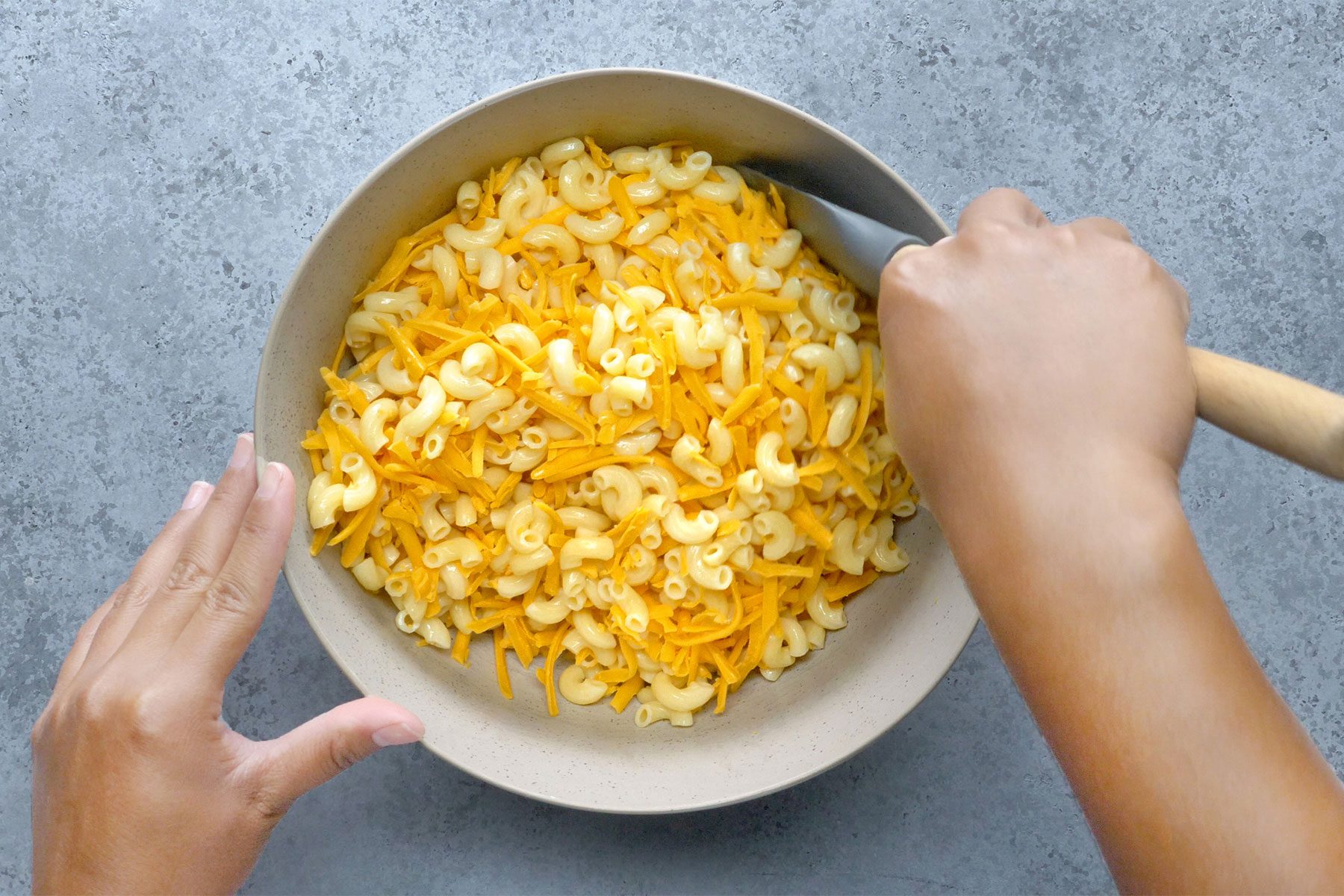 Overhead shot of drain macaroni; toss with cheddar cheese; spatula; grey marble texture background;