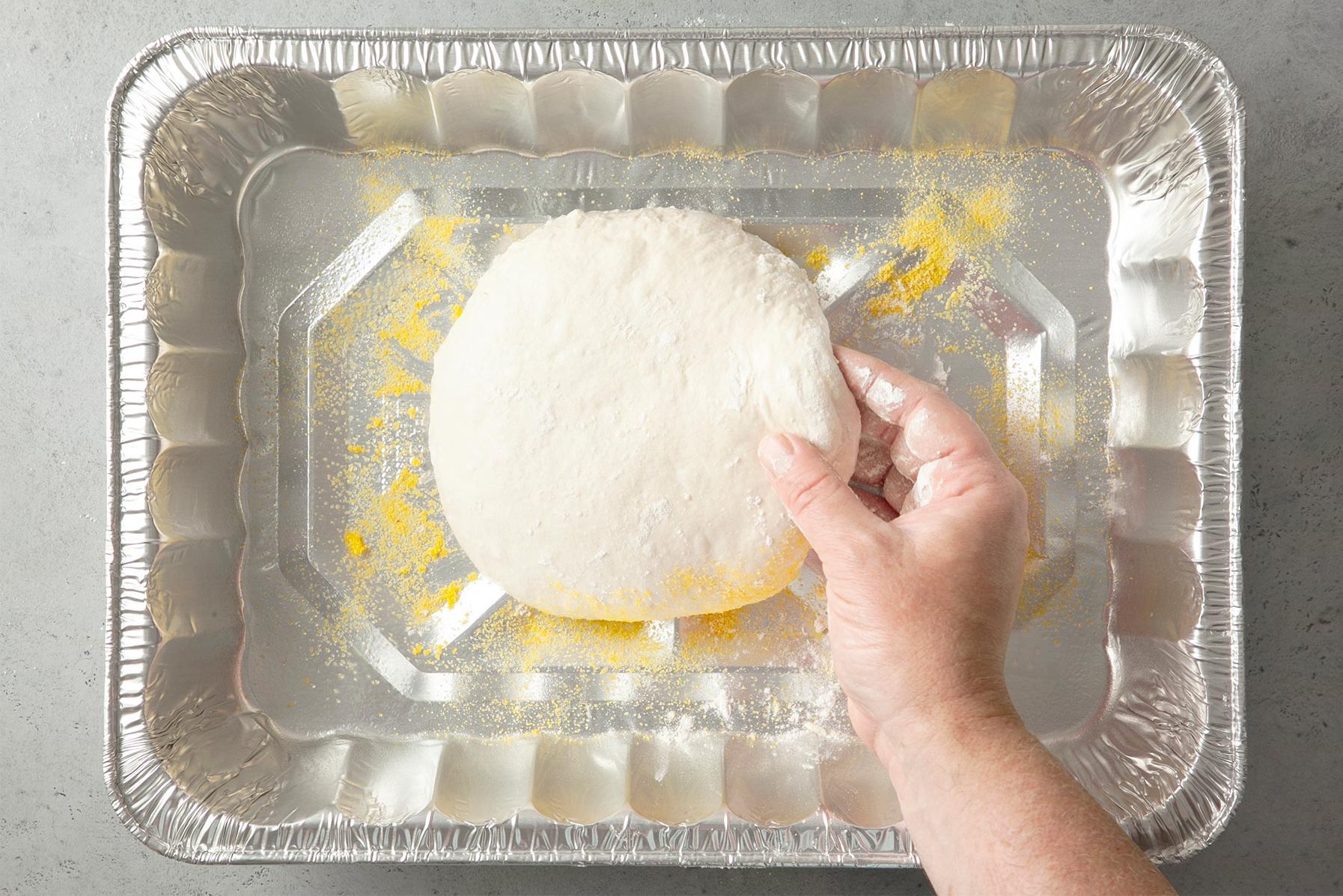 A hand holding a circular piece of pizza dough over a silver baking tray lightly dusted with flour or cornmeal. 